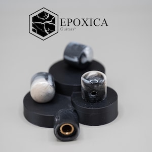 Könnte beinhalten: Eine Sammlung von Gitarrenknöpfen mit einem marmorierten Schwarz-Weiß-Design. Die Knöpfe sind zylindrisch und liegen auf schwarzen, kreisförmigen Plattformen. Der Markenname "EPOXICA Guitars" ist oben links zu sehen.