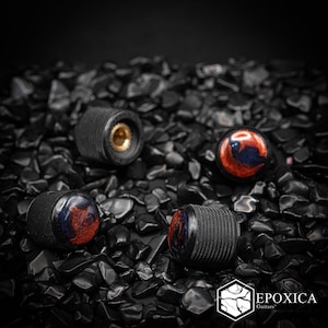 Puede incluir: Tres perillas de control de guitarra cilíndricas con un agarre moleteado negro texturizado y una parte superior lisa y pulida con un patrón de remolinos de resina roja oscura y azul oscuro. El logotipo Epoxica Guitars es visible abajo a la derecha.
