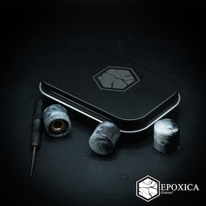 May include: Three cylindrical guitar knobs with a marbled black and white finish, featuring a textured knurled grip. A small black screwdriver is included alongside a rectangular black metal storage tin with a hexagonal logo on the lid.