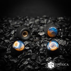 Puede incluir: Cuatro perillas de control de guitarra cilíndricas con acabado negro mate e incrustaciones de resina azul y dorada en la parte superior. Cada perilla tiene un inserto interior de color dorado metálico. El texto EPOXICA Guitars es visible en la esquina.