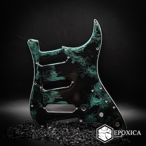 May include: A guitar pickguard featuring a black base with swirling teal and green marbled patterns. The component includes three single-coil pickup cutouts, a five-way switch slot, and three control knob holes, with the Epoxica Guitars logo visible.