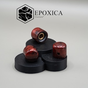 Puede incluir: Tres perillas de guitarra rojas y negras con un acabado brillante se exhiben sobre plataformas negras. Las perillas tienen forma cilíndrica con un inserto de latón. El logotipo "EPOXICA Guitars" está en el fondo.