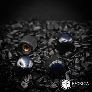 May include: A set of three cylindrical guitar control knobs with a textured black knurled side and a smooth, dark blue iridescent top. One knob is positioned to show the hollow brass interior fitting. The text EPOXICA Guitars is visible.