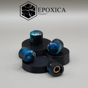 May include: Four cylindrical guitar control knobs featuring a textured black metal exterior with a marbled blue and black resin inlay on the top surface. The brand name EPOXICA Guitars is printed in black text above the knobs with a hexagonal logo.
