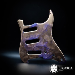 May include: A guitar pickguard featuring a marbled finish with metallic gold and deep purple swirls. The component includes three elongated cutouts for pickups and several small circular holes for mounting screws, with the text EPOXICA Guitars.