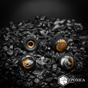 May include: Four cylindrical guitar knobs featuring a black matte exterior with a swirling amber, white, and black resin inlay on the top surface. Each knob has a textured side grip and a brass-colored metal insert. The logo Epoxica Guitars is visible.