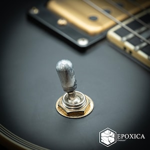 May include: A silver-colored metal toggle switch mounted on a matte black electric guitar body. The switch features a rounded tip and a hexagonal base nut. In the background, a gold-colored humbucker pickup and guitar strings are visible.