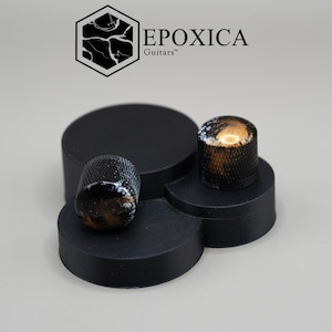 May include: Two cylindrical guitar control knobs with a textured, knurled black metal side and a marbled black, white, and metallic gold top surface. The knobs are positioned on a black, tiered, circular display stand with the text EPOXICA Guitars.