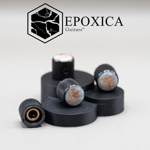 May include: A set of four black knurled guitar control knobs with white and copper-toned marbled resin tops. Each knob features a brass-colored internal splined shaft. The logo EPOXICA Guitars is displayed above the knobs in black text.
