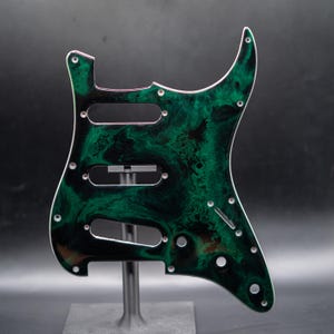 May include: A custom guitar pickguard with a swirling green and black pattern. The pickguard has three pickup cutouts, screw holes, and a white edge. It is designed to fit a Stratocaster-style guitar.
