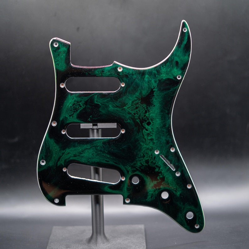 May include: A custom guitar pickguard with a swirling green and black pattern. The pickguard has three pickup cutouts, screw holes, and a white edge. It is designed to fit a Stratocaster-style guitar.
