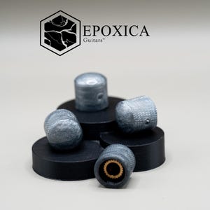 May include: A collection of silver guitar knobs with a textured surface, displayed on small black platforms. The knobs have a metallic sheen and a small hole on the side. The logo "EPOXICA Guitars" is visible in the background.