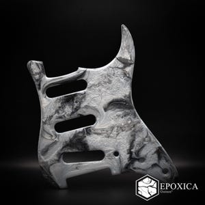 May include: A guitar pickguard featuring a marbled black and white pattern with a glossy finish. The component includes three elongated cutouts for pickups and several small circular holes for mounting screws. The logo Epoxica Guitars is visible.