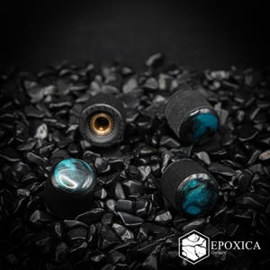 May include: Four cylindrical guitar control knobs with a textured black knurled side and a smooth, glossy top featuring a swirling teal and black resin pattern. One knob is inverted to show a brass-colored metal insert. Text reads EPOXICA Guitars.
