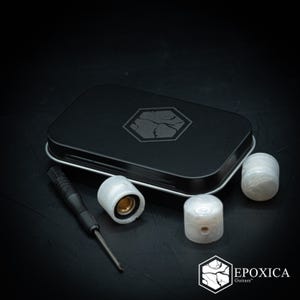 May include: A black rectangular metal tin with a hexagonal logo, three translucent white knurled guitar knobs, and a small black screwdriver. The logo features a cracked pattern, and the text Epoxica Guitars is visible in the corner.