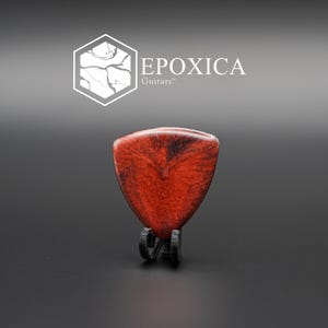 May include: A red and black guitar pick with a glossy finish, displayed on a black stand. The pick has a unique pattern and is shaped like a rounded triangle. The logo "EPOXICA Guitars" is visible in the background.