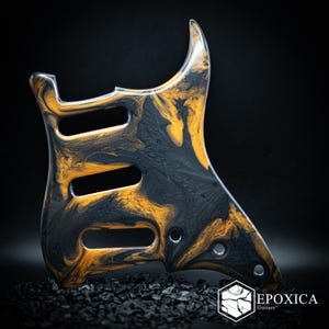 May include: A guitar pickguard featuring a marbled pattern of black and metallic gold resin. The component includes three rectangular pickup cutouts and several circular screw holes. The logo EPOXICA Guitars is printed in white in the corner.