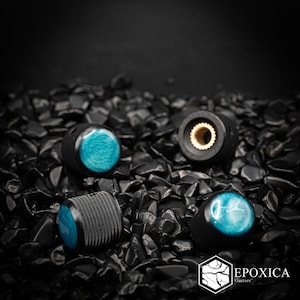 May include: Three cylindrical guitar control knobs with a black textured exterior and a vibrant teal blue circular inlay on the top. One knob is positioned to show the brass-colored interior gear mechanism. The text Epoxica Guitars is visible.