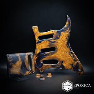 Puede incluir: Un golpeador y una placa trasera para guitarra con un patrón de mármol en espiral de color oro metálico y azul marino profundo. El conjunto incluye tres perillas de control circulares a juego y un logo que dice EPOXICA Guitars.