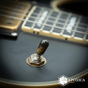 May include: A black electric guitar body featuring a toggle switch with a dark, marbled resin tip. The guitar has a matte black finish, a cream-colored binding, and a gold-toned pickup. The text EPOXICA Guitars is visible in the bottom corner.