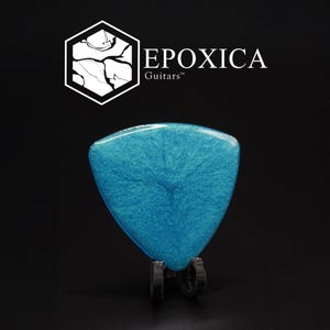 May include: A blue guitar plectrum with a shimmering, textured surface is displayed on a black stand. The plectrum is in the shape of a rounded triangle. The logo "EPOXICA Guitars" is in the upper left corner.