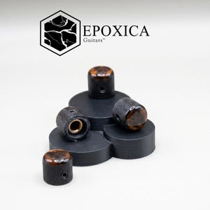 May include: A collection of guitar knobs with a tortoiseshell design. The knobs are black with a textured surface and a brass-colored interior. The knobs are arranged on a black, tiered display. The logo "EPOXICA Guitars" is visible.