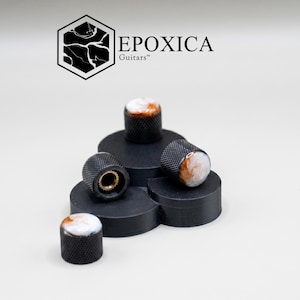 May include: A collection of black guitar knobs with a textured grip and a marbled white and brown top. The knobs are arranged on a black, tiered display, with the "EPOXICA Guitars" logo in the background.