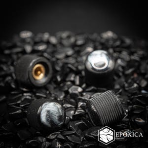 May include: Four cylindrical guitar control knobs with a textured knurled black metal side and a smooth, swirling grey and black resin top. One knob is positioned to show the hollow brass interior fitting. The logo Epoxica Guitars is visible.