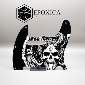 May include: Black guitar pickguard featuring a white illustration of a demonic skull with horns and a trident. The pickguard has the text "EPOXICA Guitars" and is designed for a guitar.