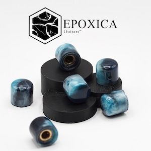 以下が含まれることがあります： 青と白の大理石模様のギターノブのコレクション。ノブは円筒形で真鍮のインサートがあり、黒いプラットフォームに配置されています。「EPOXICA Guitars」のロゴが見えます。