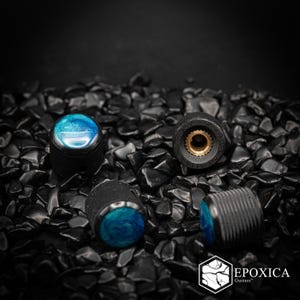 May include: Four cylindrical guitar control knobs featuring a black knurled metal exterior and a smooth, swirling blue and teal resin inlay on the top. One knob is positioned upside down to reveal a brass-colored interior gear mechanism.