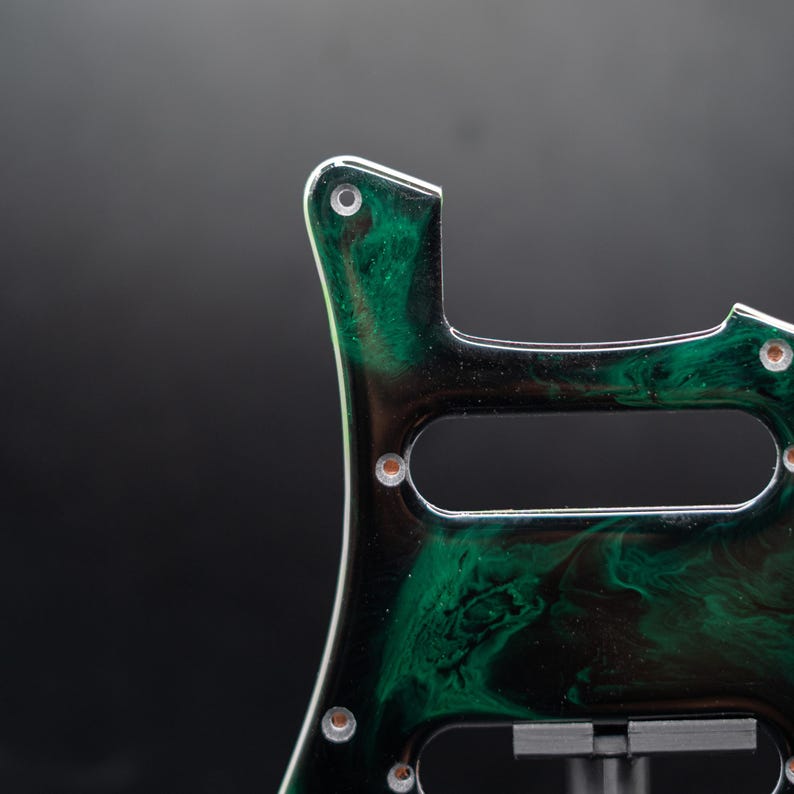 May include: Close-up of a guitar pickguard with a unique green and black marbled design. The pickguard has several screw holes and a cutout for the pickups. The edges are white, and the overall aesthetic is modern and artistic.