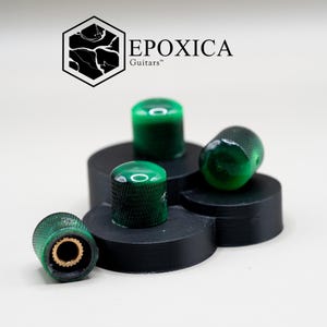 May include: Four green guitar knobs with a textured surface and gold-colored interior. The knobs are arranged on a black, multi-tiered platform. The logo "EPOXICA Guitars" is visible in the background.