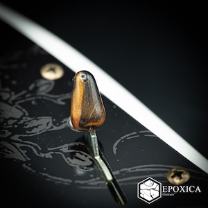 May include: A guitar pickup selector switch tip with a brown and black marbled finish, mounted on a black pickguard featuring a gray floral pattern. The pickguard includes a logo with the text EPOXICA Guitars and two visible metal screws.
