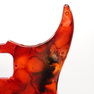 Puede incluir: Primer plano de un golpeador de guitarra con un dise&ntilde;o abstracto vibrante. El golpeador presenta una mezcla de tonos rojos, naranjas y negros, creando un efecto veteado. Tiene un acabado brillante e incluye orificios para tornillos para el montaje.