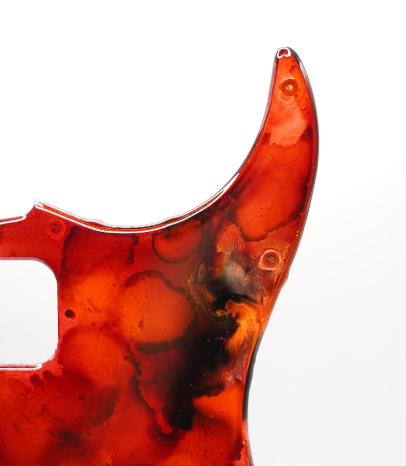 Puede incluir: Primer plano de un golpeador de guitarra con un dise&ntilde;o abstracto vibrante. El golpeador presenta una mezcla de tonos rojos, naranjas y negros, creando un efecto veteado. Tiene un acabado brillante e incluye orificios para tornillos para el montaje.