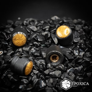 May include: Three cylindrical guitar knobs featuring a textured black knurled exterior and a circular gold-toned marbled inlay on the top. One knob is positioned to show the interior brass-colored splined shaft fitting. The text Epoxica Guitars is visible.