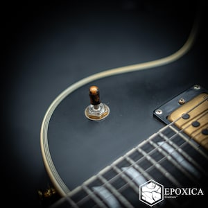 Könnte beinhalten: Ein matt-schwarzer E-Gitarrenkorpus mit cremefarbenem Binding, einem goldfarbenen Humbucker-Tonabnehmer und einem Kippschalter mit braun marmorierter Kappe. Unten rechts befindet sich ein weißes sechseckiges Logo mit dem Text Epoxica Guitars.