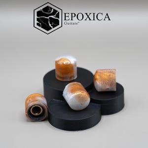 Puede incluir: Cuatro perillas de control de guitarra cilíndricas con una mezcla jaspeada de resina blanca y dorada metálica. Cada perilla tiene una superficie exterior texturizada y moleteada y un inserto interior de color latón. La marca EPOXICA Guitars es visible.