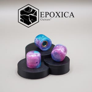 Puede incluir: Tres perillas de guitarra con un diseño veteado azul y morado se exhiben sobre una plataforma negra. Las perillas tienen forma cilíndrica con un inserto metálico. El logotipo "EPOXICA Guitars" está en el fondo.