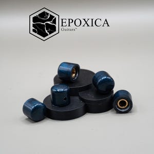 May include: A selection of blue guitar knobs with a speckled finish, displayed on black circular platforms. The knobs feature a gold metallic interior. The image also shows the "EPOXICA Guitars" logo in black.