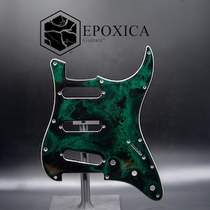 May include: A green and black guitar pickguard with a marbled design. The pickguard has three pickup cutouts and several screw holes. The brand name "EPOXICA Guitars" is printed at the top.