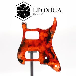 Puede incluir: Un golpeador de guitarra con un vibrante dise&ntilde;o veteado en naranja y negro. El golpeador tiene recortes para pastillas y controles, y est&aacute; montado en un soporte. El texto "EPOXICA Guitars" se muestra arriba.