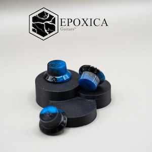 Op de afbeelding: Drie gitaarknoppen met blauw-zwarte marmerontwerpen worden gepresenteerd op een zwart platform. De knoppen hebben witte cijfermarkeringen. De merknaam "EPOXICA Guitars" staat in het zwart boven de knoppen.