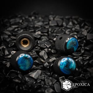 May include: Three black cylindrical guitar knobs with a textured grip and a glossy, swirling blue and black resin inlay on the top. One knob is flipped to show the brass-colored internal splined shaft. The logo EPOXICA Guitars is visible.