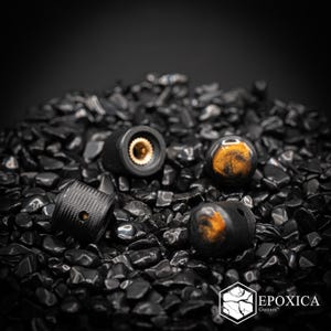 May include: Three cylindrical guitar control knobs with a textured black exterior and a metallic gold interior insert. Two knobs feature a swirling amber and black resin top, while one shows the brass-colored gear mechanism. Text reads EPOXICA Guitars.
