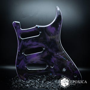 Peut inclure: Une plaque de protection pour guitare avec un motif marbré tourbillonnant de violet profond et de noir. L'élément comprend trois découpes allongées pour les micros, plusieurs petits trous de vis circulaires et une ouverture pour bouton.