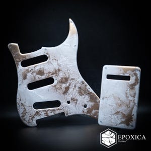 May include: A guitar pickguard and a rectangular back plate featuring a marbled white and metallic bronze finish. The pickguard has three pickup cutouts and screw holes. The brand logo EPOXICA Guitars is visible in the bottom right corner.