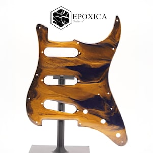 Może przedstawiać: Osłona gitary o unikalnym wzorze. Osłona ma wirujący wzór w kolorze złotym i granatowym. Posiada trzy otwory na pokrętła, gniazdo przełącznika wyboru przetwornika i kilka otworów na śruby. Widoczna jest nazwa marki "EPOXICA Guitars".