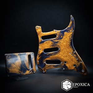 May include: A guitar pickguard and a tremolo back plate featuring a swirling marbled pattern of metallic gold and deep black resin. The pickguard has three pickup cutouts and screw holes. The logo Epoxica Guitars is visible in the corner.
